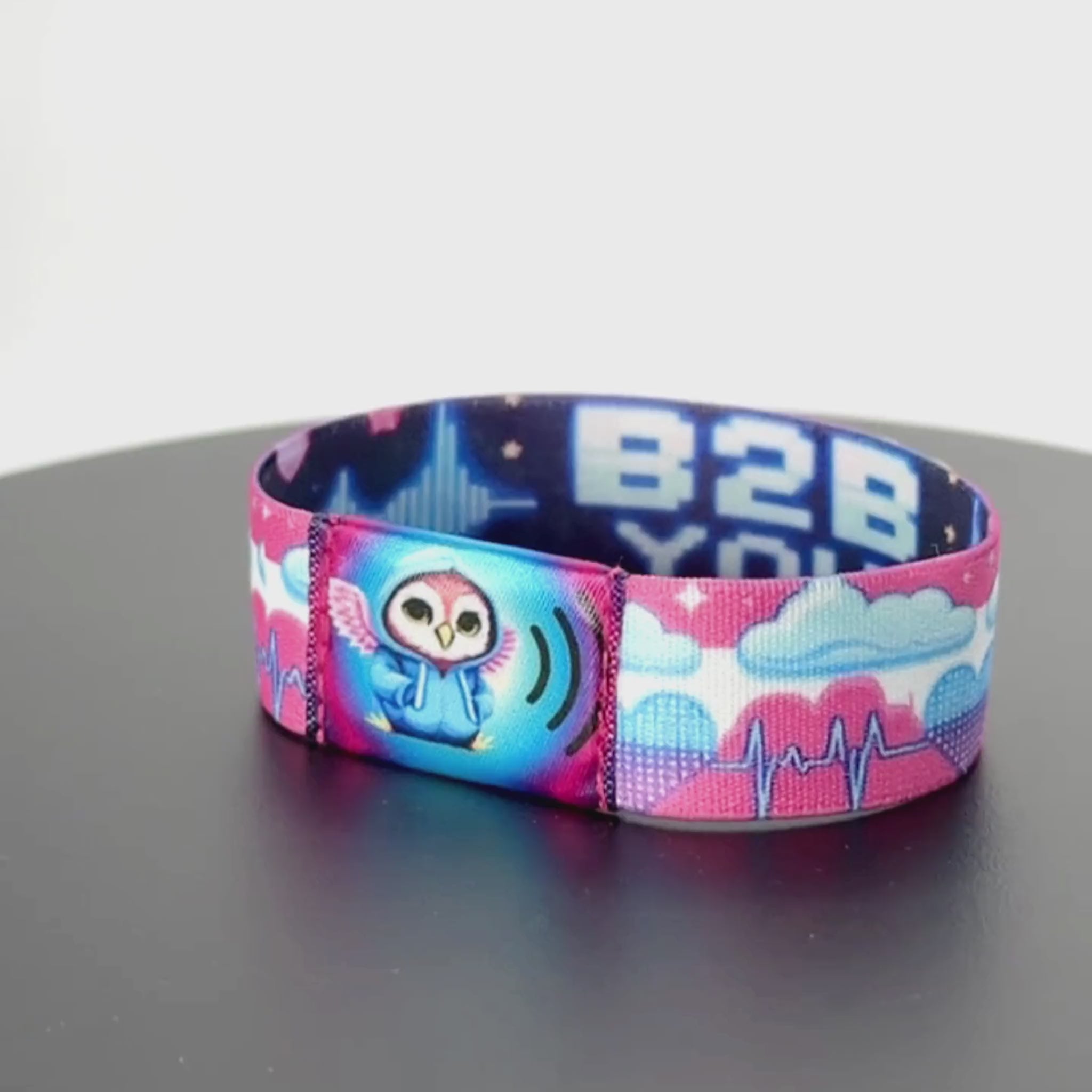 8-second rotating video of the wristband’s front, showing a pastel pink and white cloud-and-heartbeat print band with a central NFC tag featuring a cute owl in a light blue hoodie with tiny pink wings.