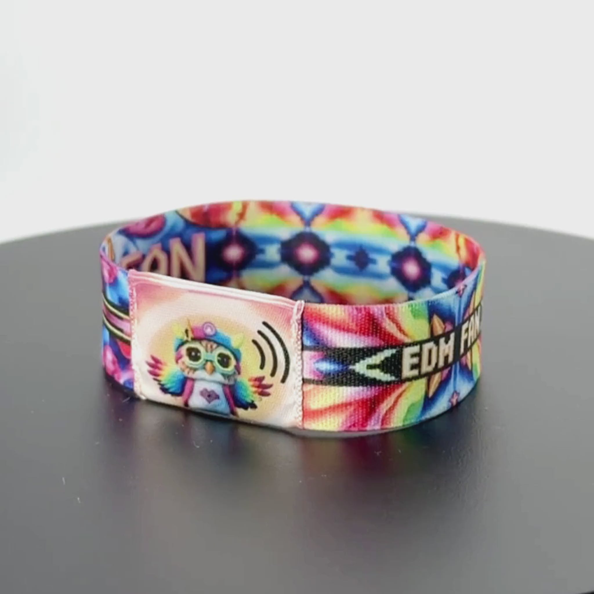 8-second rotating video of the wristband’s front, featuring a central NFC module with a pastel-colored owl wearing sunglasses and a puffer jacket, set against a vibrant rainbow tie-dye background and flanked by partial ‘FAN’ lettering.